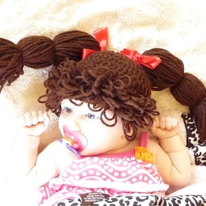 May include: A baby wearing a brown yarn wig with two pigtails tied with red bows. The wig is crocheted and has a curly texture. The baby is wearing a pink and white dress with a yellow flower.