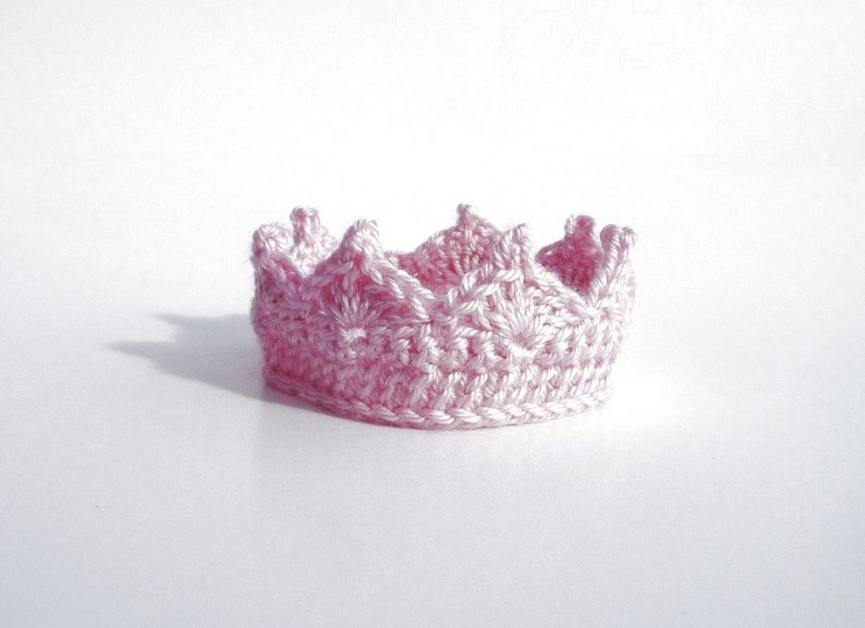 May include: A pink crocheted crown with a pointed top and scalloped edges.