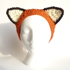 May include: A crocheted fox headband with orange, white, and black yarn. The headband is on a white mannequin head.