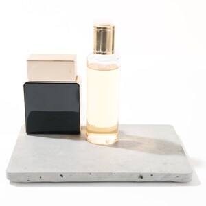 Peut inclure: Deux flacons de parfum sur un plateau en b&eacute;ton gris. Le flacon de gauche est noir avec un dessus dor&eacute;. Le flacon de droite est en verre transparent avec un dessus dor&eacute; et un liquide jaune clair &agrave; l'int&eacute;rieur.