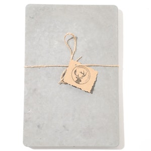 Peut inclure: Une planche &agrave; d&eacute;couper en b&eacute;ton gris avec une &eacute;tiquette en papier brun attach&eacute;e avec de la ficelle. L'&eacute;tiquette porte un logo avec une t&ecirc;te de cerf et le texte "Cedarwood Creek Co."