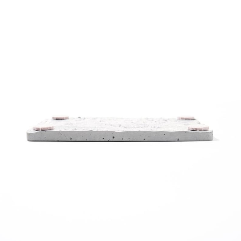 Peut inclure: Un dessous de verre carr&eacute; en b&eacute;ton avec une surface rugueuse et textur&eacute;e. Le dessous de verre a quatre petits pieds en caoutchouc rose sur le dessous.