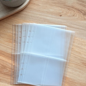 Könnte beinhalten: Transparente Kunststoff-Ordnerhüllen mit mehreren Fächern, zum Aufbewahren von Fotos oder Dokumenten. Die Hüllen sind auf einer hellen Holzmaserung gestapelt, mit einer kleinen Pflanze im Hintergrund.