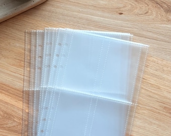 May include: Clear plastic binder sleeves with multiple pockets, designed for storing photos or documents. The sleeves are stacked on a light wood-grain surface, with a small plant in a gray pot visible in the background.