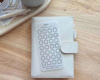 May include: A white leather planner with a snap closure, holding a clear plastic tracker card. The card has a grid of house icons and text that reads "RENT" and "THE MILLENNIAL DOLLAR". The planner is on a wooden surface.
