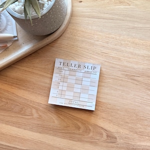 Puede incluir: Un pequeño resguardo de cajero cuadrado con el texto "TELLER SLIP" en la parte superior. El resguardo tiene columnas para billete, cantidad y cantidad, con denominaciones de 1 a 100. Las palabras "TOTAL" y "THE MILLENNIAL DOLLAR" también están impresas.