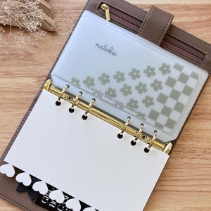 May include: A brown faux leather planner with gold rings and a zip closure. The planner features a clear pocket labelled "matcha" with a floral and chequered design. A white page with a heart-shaped border is visible.