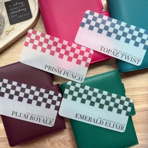 Könnte beinhalten: Eine Sammlung farbenfroher Geldbörsen und Karten. Die Karten zeigen Schachbrettmuster in verschiedenen Farben, darunter Rot, Blau und Grün, mit Texten wie "Prism Punch" und "Topaz Twist". Die Geldbörsen sind in den Farbtönen Pink, Türkis, Burgunderrot und Grün gehalten.