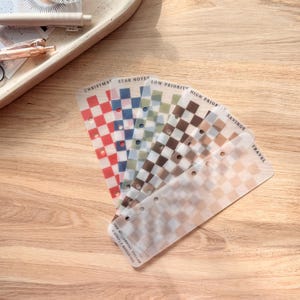 May include: A collection of translucent planner dividers with a checkered pattern in various colors, including red, blue, green, brown, and beige. Each divider is labeled with text such as "Christmas," "Star Notes," and "Savings."
