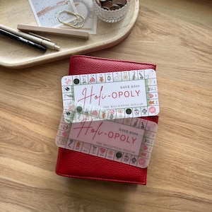 Puede incluir: Una cartera de cuero roja con dos superposiciones transparentes con el texto "Holi-OPOLY" y gráficos de tablero de juego. Las superposiciones dicen "Ahorra $300" y "The Millennial Dollar". La cartera está sobre una superficie de madera.