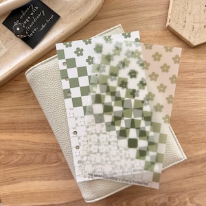 Könnte beinhalten: Ein cremefarbener Planer mit drei dekorativen Einlagen. Die Einlagen weisen ein grün-weißes Karomuster, florale Designs und den Text "The Millennial Dollar x Simply Budgeted" auf. Ein kleines schwarzes Etikett mit weißem Text ist ebenfalls sichtbar.