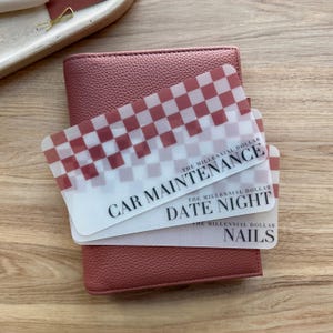 May include: A dusty rose leather wallet with three translucent cards. The cards have a red and white checkered pattern and the words "CAR MAINTENANCE", "DATE NIGHT", and "NAILS" printed on them. The text "THE MILLENNIAL DOLLAR" is also printed on the cards.