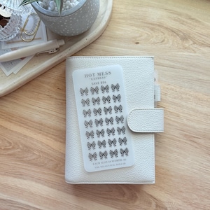 May include: A white leather planner with a snap closure, holding a clear sticker sheet. The sticker sheet features black and white checkered bow icons and text that reads "HOT MESS EXPRESS SAVE $56". The planner is on a wooden surface.