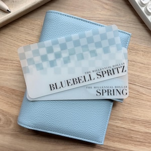 Puede incluir: Una cartera de cuero azul claro con superficie texturizada sostiene dos tarjetas translúcidas. Las tarjetas presentan un patrón de cuadros y el texto "BLUEBELL SPRITZ" y "SPRING". La cartera y las tarjetas están sobre una superficie de madera.