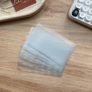 May include: A stack of clear, plastic binder sleeves with the text "THE MILLENNIAL DOLLAR" printed on the side. The sleeves are on a wooden surface, with a calculator and a decorative tray in the background.