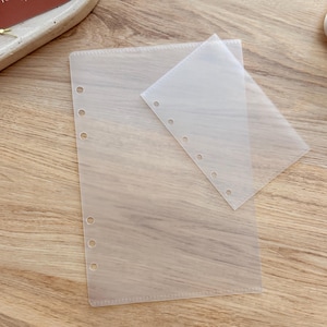 Pode incluir: Duas folhas de plástico transparentes e translúcidas para encadernação com vários orifícios pré-perfurados para organização. Uma folha é maior e a outra menor, ambas retangulares. As folhas estão sobre uma superfície de madeira castanha clara.