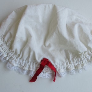 Puede incluir: Bonnet de algodón blanco con ribete de encaje y una cinta roja atada en la parte inferior.
