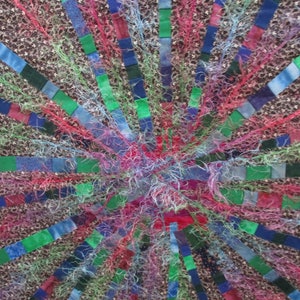 May include: A close-up of a colourful quilt with a radial design. The quilt is made of various fabrics in shades of blue, green, red, and pink. The fabric is arranged in a circular pattern, with the centre of the circle being the focal point. The quilt is made of many small squares of fabric, which are sewn together to create a larger design.