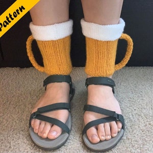 Könnte beinhalten: Ein Paar gestrickte Bierkrüge-Beinwärmer in Gelb und Weiß. Die Beinwärmer werden über einem Paar dunkelgrüner Sandalen getragen.