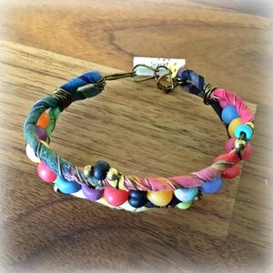 May include: A colorful bracelet with a fabric band wrapped in wire. The bracelet features a variety of beads in shades of red, blue, yellow, and purple. The clasp is made of metal.
