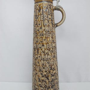 May include: A tall, cylindrical ceramic beer stein with a mottled brown and tan glaze. The exterior features embossed German text covering the surface. It includes a pewter lid with a thumb-operated hinge and a matching curved handle on the side.