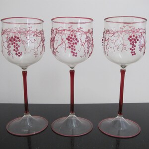 May include: Three clear glass wine glasses with a red rim and stem. The glasses are decorated with a grapevine design in pink.