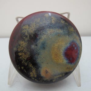 Peut inclure: Un objet rond en céramique émaillée, avec un extérieur marron foncé et un motif central aux teintes rouge, jaune, bleu et noir. L'objet est présenté sur un support transparent, mettant en valeur sa palette de couleurs et sa texture uniques.