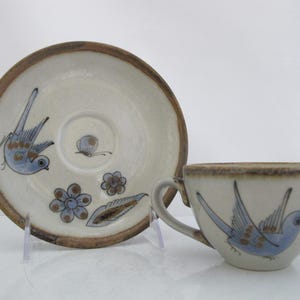 May include: A ceramic teacup and matching saucer set featuring a hand-painted light blue bird, small blue flowers, and a butterfly motif on an off-white background with a brown speckled rim on both the cup and the saucer edges.