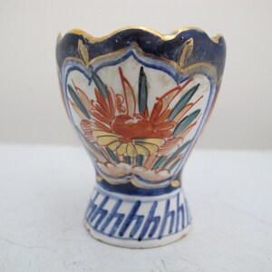 Può includere: Un piccolo vaso in ceramica bianca con un motivo floreale blu e oro. Il vaso ha un bordo a festoni e una base svasata.