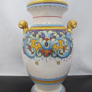 May include: A white ceramic vase with a bulbous body and two gold handles. The vase features intricate hand-painted floral and scrollwork designs in blue, yellow, and red. The base and rim are adorned with colourful bands.