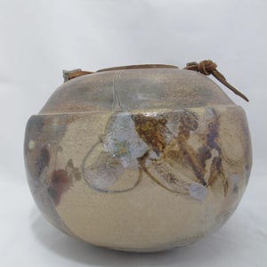May include: A handmade ceramic jar with a rounded form and a lid. The jar has a beige base with brown and blue abstract patterns. It features a brown leather handle attached to the lid. The jar is suitable for storage or display.