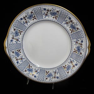 Puede incluir: Un plato para pastel de cerámica blanca con un patrón floral azul y dorado. El plato tiene un borde festoneado y dos asas.