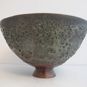 May include: A ceramic bowl with a textured, pitted surface. The bowl has a dark, earthy colour palette with shades of brown, green, and black. The bowl has a rounded shape and a small, pedestal base.