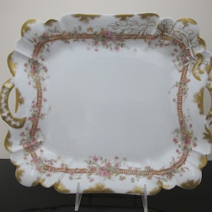 Puede incluir: Una bandeja de servir de porcelana blanca con ribetes dorados y diseños florales rosas. La bandeja tiene dos asas y un borde festoneado.
