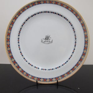 Puede incluir: Plato de cerámica blanca con un borde decorativo con diamantes rojos, azules y naranjas. El borde tiene una banda dorada. Un escudo de armas está centrado en el plato. Este plato es adecuado para servir alimentos.