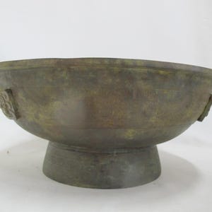 Può includere: Una ciotola in stile antico, color bronzo, con un bordo ampio e arrotondato e una base a piedistallo. La ciotola presenta manici decorativi su entrambi i lati e una superficie strutturata, che suggerisce età e maestria. La ciotola è probabilmente in metallo.