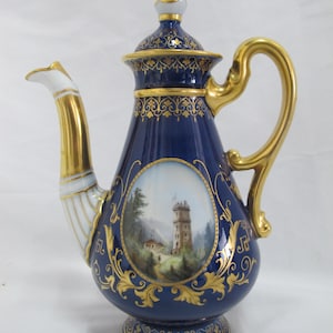 May include: A cobalt blue porcelain teapot with gold accents. The teapot features a hand-painted landscape scene within an oval frame. The handle, spout, and lid are gold-plated. Intricate gold filigree decorates the body and base of the teapot.