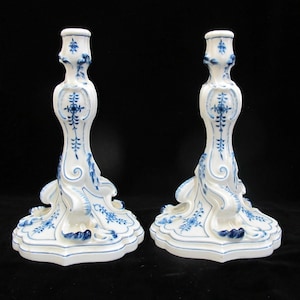 May include: Two white ceramic candlesticks with blue floral patterns. The candlesticks have a flared base, a slender body, and a top designed to hold a candle. The blue and white design is a classic style.