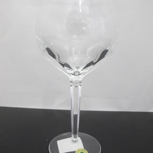 Puede incluir: Una copa de vino de cristal transparente con un cuenco facetado y un tallo largo y delgado. La copa se asienta sobre una base redonda con una pequeña pegatina. La copa está diseñada para mejorar la experiencia de beber vino.