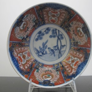 May include: A decorative ceramic plate with a detailed design. The plate features a central image of a bird perched on a branch, surrounded by floral and geometric patterns in red, blue, and orange. The plate is likely vintage.