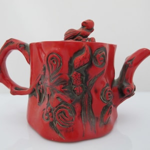 Puede incluir: Una tetera de cerámica de color rojo brillante con una forma orgánica texturizada que recuerda a un tocón de árbol. La superficie presenta patrones oscuros en espiral y tallas en forma de hoja. Una pequeña figura de pájaro esculpida se asienta sobre la tapa, y el asa y el pico tienen forma de ramas nudosas.