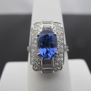 May include: A silver ring with a large oval blue gemstone at the center, surrounded by smaller clear stones. The ring has a rectangular design with additional clear stones and a band.