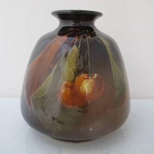 May include: A dark brown ceramic vase with a glossy finish. The vase features a hand-painted design of two orange cherries with green leaves. The vase has a rounded body and a narrow neck.