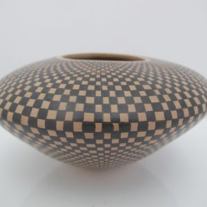 May include: A low-profile, wide-mouthed ceramic bowl featuring a repeating black and tan checkerboard pattern that wraps around the entire exterior surface. The vessel has a rounded, tapered base and a smooth, matte finish across its curved form.