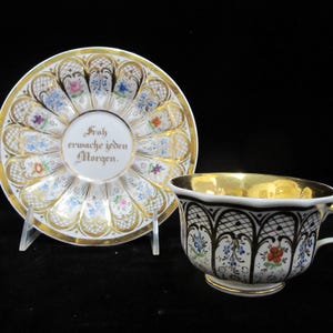 Könnte beinhalten: Ein Vintage-Tee-Set mit Tasse und Untertasse. Die Tasse hat einen goldenen Rand und Henkel, mit einem schwarz-weißen Gittermuster und floralen Akzenten. Die Untertasse hat ein Blumen- und Goldmuster mit dem Text "Froh erwache jeden Morgen."