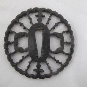 Peut inclure: Une tsuba en métal sombre, une garde de sabre japonaise, avec un design ajouré complexe. La garde présente une ouverture ovale centrale et des motifs floraux et d'oiseaux détaillés. La couleur sombre et le savoir-faire de la tsuba suggèrent une pièce ancienne.