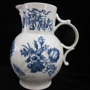 Puede incluir: Una jarra de porcelana blanca con un diseño floral azul. La jarra tiene un cuerpo redondo y un asa curva. El diseño floral incluye rosas, hojas y una pera.