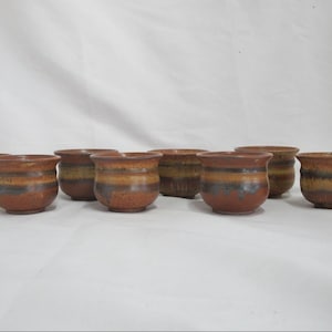 Puede incluir: Un conjunto de nueve pequeñas tazas de cerámica. Cada taza tiene forma cilíndrica con un borde ligeramente ensanchado. Están decoradas con bandas horizontales de esmalte marrón, beige y verde azulado oscuro. Se muestran sobre una superficie blanca.