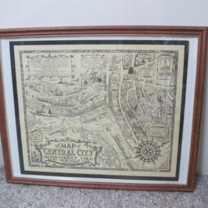 May include: A framed black and white map of Central City, Gilpin County, Colorado. The map is detailed and includes many streets and buildings. The map is surrounded by a black border and a brown frame.