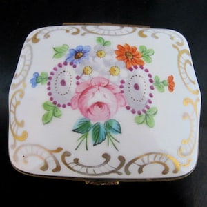 May include: A white porcelain trinket box with a hinged lid. The lid is decorated with a floral design featuring pink, red, yellow, blue, and green flowers. The design is surrounded by a gold-colored border.